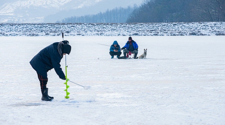 A person using an ice auger with two people ice fishing in the background.