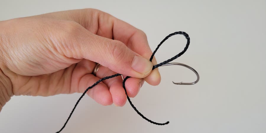 A hand holding fishing line looped above a hook's shaft against a white background.